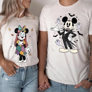 Könnte beinhalten: Zwei weiße T-Shirts mit Halloween-Motiven. Das linke T-Shirt zeigt eine Mausfigur in einem Patchwork-Kleid mit Schleife. Das rechte T-Shirt zeigt eine Mausfigur in einem Nadelstreifenanzug, umgeben von Geistern und Fledermäusen.