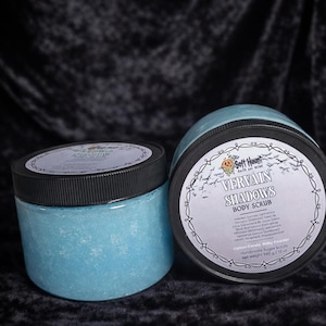 May include: Two jars of blue body scrub with black lids. The jars are labeled "Vervain Shadows Body Scrub" and have a decorative label with a floral design. The scrub has a light blue color and a granular texture. The net weight is 12 oz.
