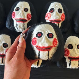May include: A collection of black, white, and red novelty items shaped like a character's face from a horror movie. The faces have black hair, white skin, red cheeks, and red lips. The items are arranged on a black surface.