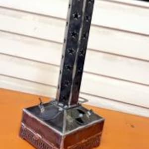 Könnte beinhalten: Ein handgefertigter Kerzenhalter aus Metall mit quadratischer Basis und hohem, rechteckigem Turm. Der Turm hat Sternaussparungen, und die Basis hat dekorative Kanten. Der Kerzenhalter ist aus dunklem Metall gefertigt und steht auf einer orangefarbenen Oberfläche.