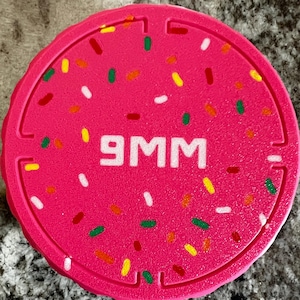 May include: A circular, bright pink rubber cover featuring the text 9MM in white, centered on the surface. The design is decorated with small, scattered, multi-colored sprinkles in shades of red, yellow, green, and white across the top.