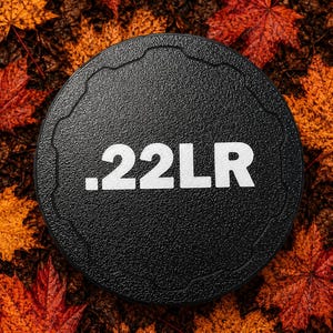 May include: A circular black textured cap featuring the white text .22LR printed in the center. The cap has a scalloped edge design and a matte finish, providing a distinct contrast against the surrounding vibrant orange and red autumn leaves.