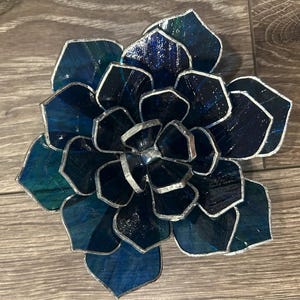 May include: A handcrafted stained glass succulent in shades of blue and teal. The glass petals are outlined with silver solder, creating a dimensional effect. This decorative piece is designed to resemble a succulent plant.