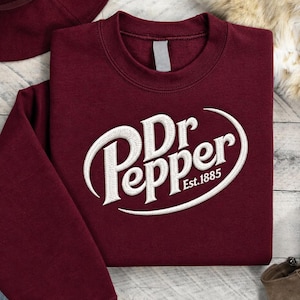 May include: A maroon long-sleeve sweatshirt featuring the white embroidered Dr Pepper logo with the text Est. 1885 centered on the chest. The garment has a crew neckline and long sleeves, presented as a flat lay item of clothing.