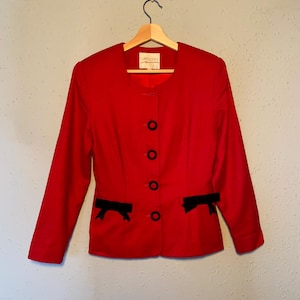 May include: A bright red blazer with long sleeves, black buttons, and black bow accents on the pockets. The blazer is hanging on a wooden hanger. The label reads "CAPUCCIN".