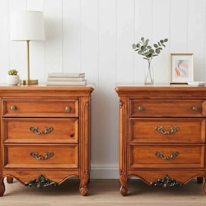 Może przedstawiać: Dwa pasujące drewniane stoliki nocne, każdy z trzema szufladami. Stoliki nocne mają ozdobne detale, w tym dekoracyjne uchwyty szuflad i rzeźbione akcenty. Lampa, książki i przedmioty dekoracyjne stoją na jednym ze stolików.