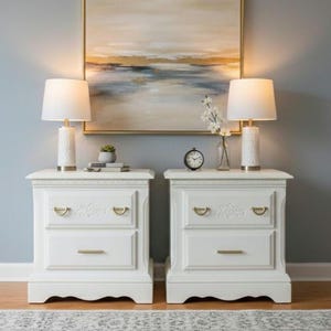 May include: Two white bedside tables with gold hardware and lamps with white shades. A painting with gold and blue tones hangs above the tables. A small clock and vase with flowers are on one table.