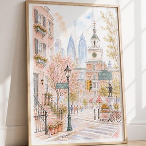 Könnte beinhalten: Ein gerahmter Aquarelldruck einer Straßenszene in Philadelphia mit rosa Kirschblütenbäumen, einem historischen Backsteingebäude mit Uhrturm, einer Straßenlaterne, einem Brezelwagen mit der Aufschrift Philly Pretzels und Wolkenkratzern.