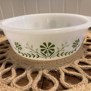 Puede incluir: Fuente ovalada blanca con diseño floral verde. El plato tiene dos asas y está sobre un salvamanteles tejido. El patrón floral incluye flores y hojas estilizadas.