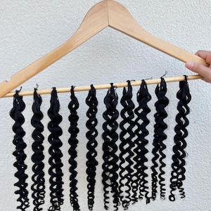 May include: A wooden clothes hanger displaying multiple strands of black curly hair extensions. The extensions are attached to a wooden dowel and hang in a row. The background is a plain white wall.