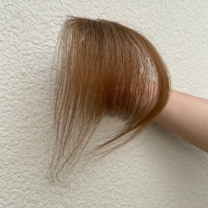 May include: A light brown hair piece, possibly a fringe or a bang extension, against a plain background. The hair is straight and has a warm, natural colour. The piece is likely designed to add volume or change the hairstyle.