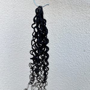May include: A bundle of black, curly hair extensions hanging from a silver metal hook. The hair is tightly curled and appears to be synthetic. The background is a plain, off-white surface.