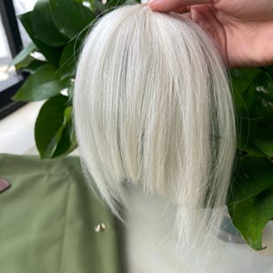 May include: A clip-in hair extension piece featuring straight, bright white synthetic fibers. The hair is styled with a blunt, straight-cut fringe that falls to a medium length, designed to be attached to the top of the head for added volume.