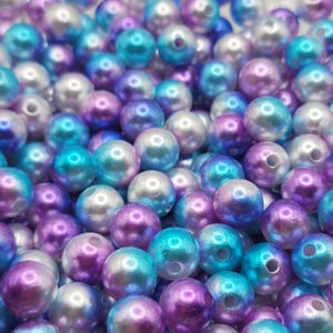 May include: A close-up shot of a pile of iridescent beads in shades of purple, teal, and white. The beads are round with a glossy finish and a small hole through the center. These beads are suitable for jewelry making and crafts.