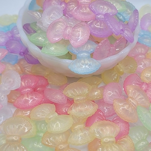 May include: A collection of pastel-colored resin bow charms in various shades of pink, yellow, blue, green, and purple. Some charms are in a white bowl, while others are scattered around. These decorative bows are ideal for crafting and embellishments.
