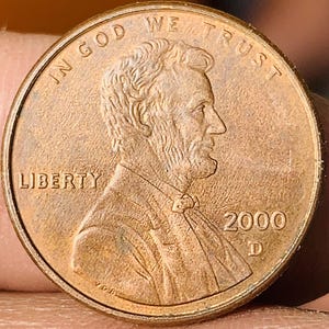 Puede incluir: Primer plano de un centavo estadounidense de 2000 de color cobre. La moneda presenta el perfil de Abraham Lincoln, las palabras "IN GOD WE TRUST" y "LIBERTY", y el año 2000.
