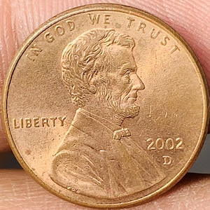 May include: A copper-colored 2002 United States penny. The obverse features a profile of Abraham Lincoln, the word "LIBERTY", and the inscription "IN GOD WE TRUST". The year "2002" and the mint mark "D" are also visible.