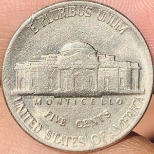 May include: A silver United States nickel coin featuring the Monticello building. The coin has the text "E PLURIBUS UNUM", "UNITED STATES OF AMERICA", "MONTICELLO", and "FIVE CENTS".
