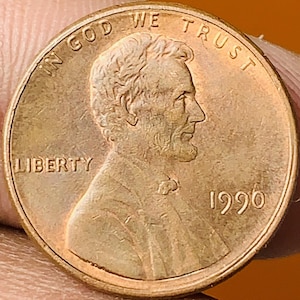 May include: A copper-colored 1990 Lincoln penny. The obverse features a profile of Abraham Lincoln, the word "LIBERTY", and the phrase "IN GOD WE TRUST". The year "1990" is also visible.