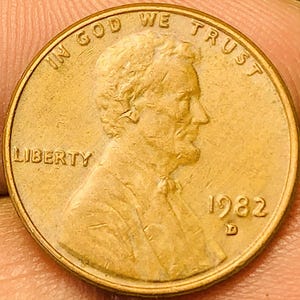 Puede incluir: Primer plano de una moneda Lincoln de 1982, de color cobre. La moneda presenta el perfil de Abraham Lincoln, la palabra "LIBERTY" y la frase "IN GOD WE TRUST". El año "1982" es visible.