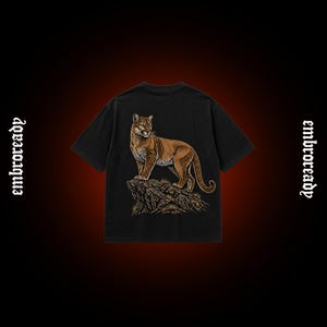 Pode incluir: Uma t-shirt preta de manga curta com um gr&aacute;fico detalhado de um puma castanho em cima de uma forma&ccedil;&atilde;o rochosa. A palavra "embroready" est&aacute; impressa numa fonte de estilo g&oacute;tico nos lados esquerdo e direito do design da t-shirt.