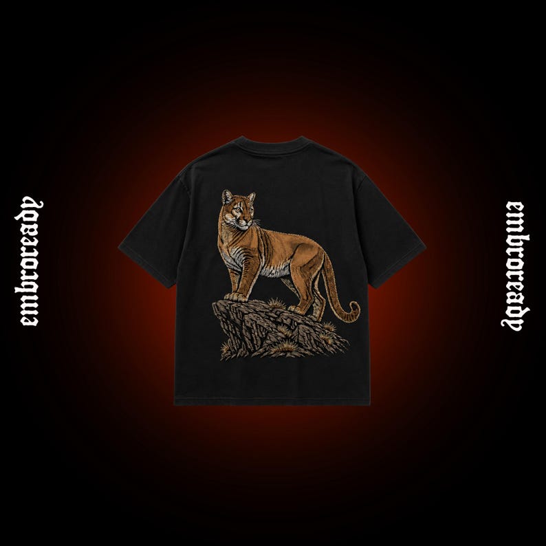 Pode incluir: Uma t-shirt preta de manga curta com um gr&aacute;fico detalhado de um puma castanho em cima de uma forma&ccedil;&atilde;o rochosa. A palavra "embroready" est&aacute; impressa numa fonte de estilo g&oacute;tico nos lados esquerdo e direito do design da t-shirt.