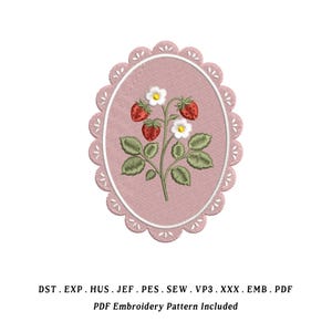 Może przedstawiać: Wyszywany owalny wzór przedstawiający roślinę truskawki z czerwonymi jagodami, białymi kwiatami i zielonymi liśćmi. Wzór jest umieszczony na różowym tle i obramowany falistą krawędzią. Poniżej znajduje się tekst "PDF Embroidery Pattern Included".