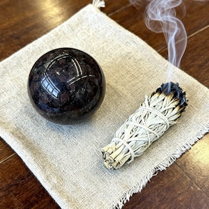 Puede incluir: Una esfera de color marrón oscuro pulida con una superficie lisa y reflectante junto a un atado de salvia humeante. El atado de salvia está envuelto en hilo blanco y emite un fino rastro de humo blanco, sobre un trozo de tela beige cuadrada.