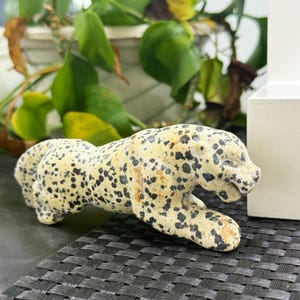 May include: A carved stone figurine of a crouching leopard with a cream-coloured base and irregular black spots. The sculpture depicts the animal in a low, prowling stance with its head extended forward and mouth slightly open, showing carved details.