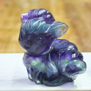 Può includere: Una statuetta in fluorite intagliata a forma di volpe con grandi orecchie a punta e una coda arricciata. La pietra presenta una superficie lucida e traslucida con bande di colore viola intenso e verde su tutto il corpo della scultura animale.