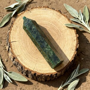 May include: A polished, hexagonal dark green crystal point with mottled textures and a pointed tip. The stone features deep forest green hues with subtle black inclusions throughout its elongated, faceted structure, displaying a smooth, reflective surface.
