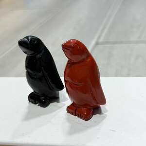 Puede incluir: Dos pequeñas figuras de piedra lisa tallada con forma de pingüinos. Una figura es de color negro sólido con una superficie pulida y reflectante, y la otra es de un rojo sólido profundo con un acabado liso y brillante, ambas de pie.