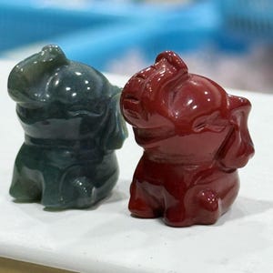 May include: Two small carved stone elephant figurines. One elephant is a deep, dark green colour with a smooth, polished finish, while the other is a solid, vibrant red colour with a similar glossy, reflective surface texture.