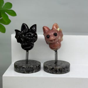 May include: Two carved stone bat figurines mounted on metal springs and circular granite bases. One bat is solid black, while the other is a mottled pinkish-brown stone. Both figures feature large, dark, rounded eyes and pointed ears.