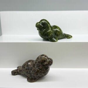 Può includere: Due piccole statuette di animali a forma di foca. Una statuetta è in pietra lucida verde scuro con una sottile venatura. La seconda statuetta è in pietra marrone caratterizzata da un motivo distinto di macchie circolari più scure.