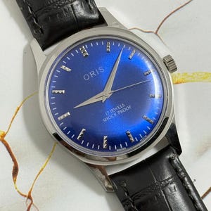 May include: A vintage Oris wristwatch with a vibrant blue dial and silver hands and markers. The watch features a stainless steel case and a black leather band. The dial reads "17 Jewels Shock Proof" and "Swiss Made".