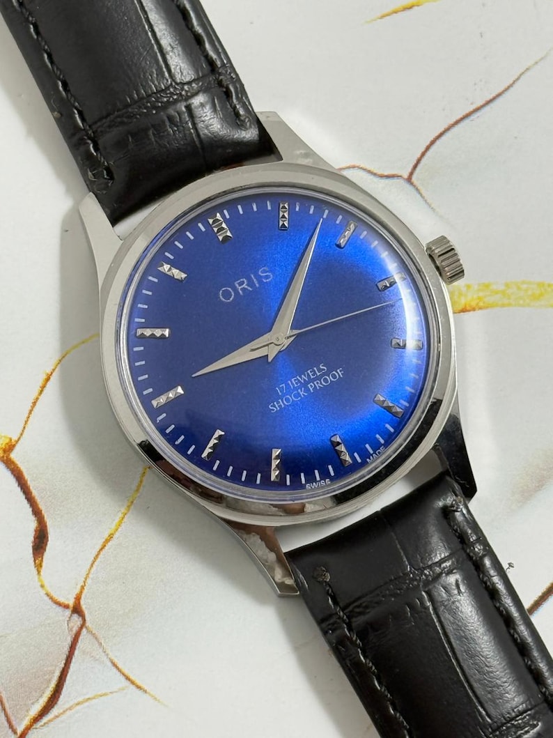 May include: A vintage Oris wristwatch with a vibrant blue dial and silver hands and markers. The watch features a stainless steel case and a black leather band. The dial reads "17 Jewels Shock Proof" and "Swiss Made".