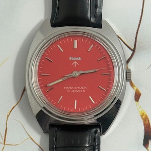 May include: A vintage wristwatch with a red face and silver hands. The watch has a silver case and a black leather band. The face includes the text "Para Shock 17 Jewels".