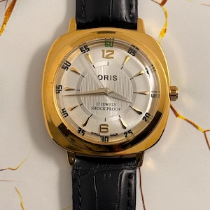 May include: A gold-tone wristwatch with a square-shaped case and a black leather strap. The silver dial features a vertical striped pattern, gold-tone hour markers, the text ORIS, 17 JEWELS, SHOCK PROOF, and SWISS MADE, with gold hands and a crown.