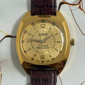 May include: A gold-toned Oris wristwatch with a brown leather band. The watch face features the text "ORIS", "17 JEWELS", and "SHOCK PROOF". The watch has a gold-colored case and a textured, circular dial with hour markers.