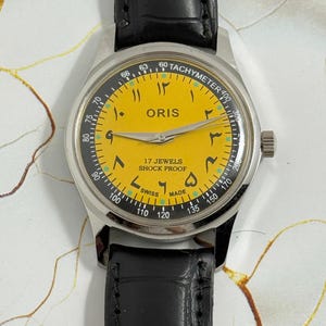 May include: A vintage Oris wristwatch with a yellow dial and Arabic numerals. The watch features a silver-tone case, a black leather strap, and a tachymeter bezel. The dial reads "17 Jewels Shock Proof Swiss Made".
