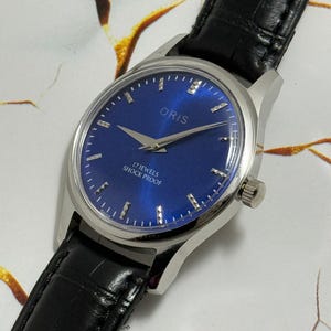 May include: A close-up of an Oris wristwatch with a blue face and silver casing. The watch has a black leather band. The watch face displays the brand name "ORIS" and the text "17 JEWELS SHOCK PROOF".
