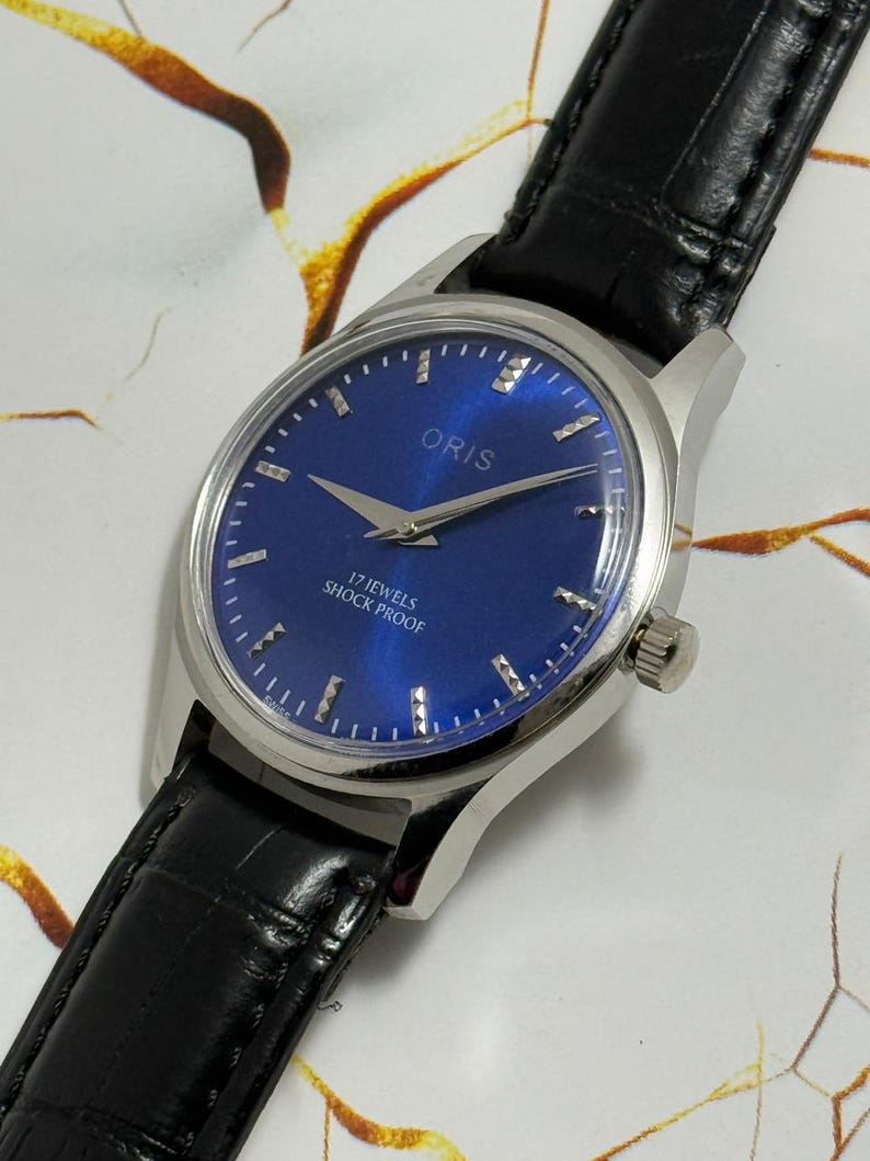May include: A close-up of an Oris wristwatch with a blue face and silver casing. The watch has a black leather band. The watch face displays the brand name "ORIS" and the text "17 JEWELS SHOCK PROOF".