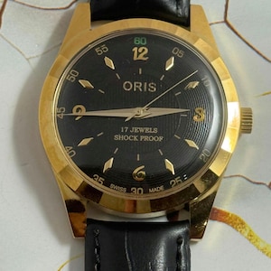 Peut inclure: Une montre-bracelet Oris dorée avec un cadran noir, des aiguilles et des index dorés. La montre a un bracelet en cuir noir et indique "17 Jewels Shock Proof". La montre est de style vintage.