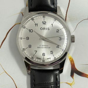 以下が含まれることがあります： 黒い革バンドが付いたシルバーカラーのORIS腕時計。文字盤はシルバーで、黒い数字と針があります。「ORIS」、「17 JEWELS」、「SHOCK PROOF」、「SWISS MADE」の文字が見えます。時計は丸い文字盤とリューズを備えています。
