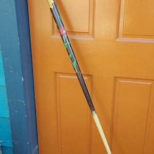 May include: A wooden walking stick with a black handle and a brown shaft. The shaft is decorated with painted flowers in shades of yellow, red, and green.