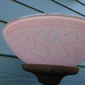 Puede incluir: Una pantalla de lámpara de cristal rosa vintage con diseños florales y de volutas. La pantalla tiene un borde decorativo con cuentas y está montada sobre una base oscura y ornamentada. El fondo es una superficie de madera azul.