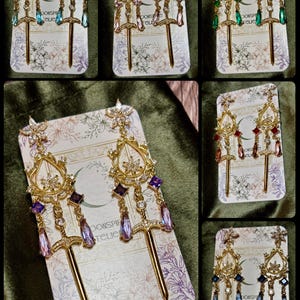 May include: Gold-toned drop earrings with ornate filigree and jewel accents. Each earring features a sword-shaped charm and dangling crystals in various colors, including blue, pink, and green. The earrings are displayed on floral-patterned cards.