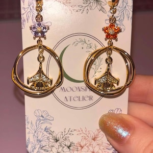 May include: A pair of gold-toned hoop earrings featuring a central jet aircraft charm encrusted with clear stones. Each earring is topped with a small flower-shaped accent, one in light purple and one in orange, attached to a display card labeled MOONSPARK ATELIER.