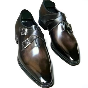 Op de afbeelding: Een paar bruin lederen geklede schoenen met een glanzende afwerking. De schoenen hebben een dubbel monk strap design met zilveren gespen en een spitse neus. De schoenen worden op een witte achtergrond gepresenteerd.